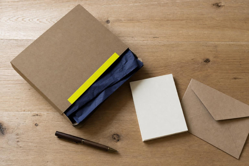 Caja plana de cartón natural con precinto amarillo, una tarjeta en blanco y un sobre, vistos desde arriba sobre una mesa de roble.